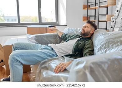 Moving, People And Real Estate Concept - Tired Man With Boxes Resting On Sofa Covered With Plastic Sheeting At New Home