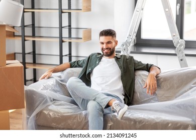 Moving, People And Real Estate Concept - Happy Smiling Man With Boxes Resting On Sofa Covered With Plastic Sheeting At New Home