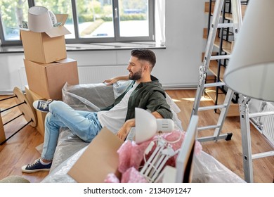 Moving, People And Real Estate Concept - Happy Smiling Man With Boxes Resting On Sofa Covered With Plastic Sheeting At New Home