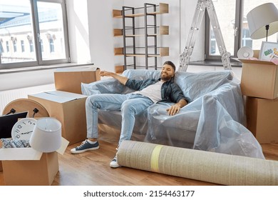 Moving, People And Real Estate Concept - Happy Smiling Man With Boxes Resting On Sofa Covered With Plastic Sheeting At New Home