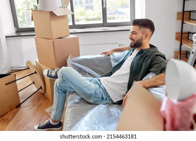 Moving, People And Real Estate Concept - Happy Smiling Man With Boxes Resting On Sofa Covered With Plastic Sheeting At New Home