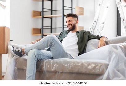 Moving, People And Real Estate Concept - Happy Smiling Man With Boxes Resting On Sofa Covered With Plastic Sheeting At New Home