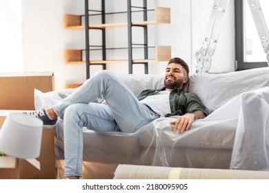 Moving, People And Real Estate Concept - Happy Smiling Man With Boxes Resting On Sofa Covered With Plastic Sheeting At New Home