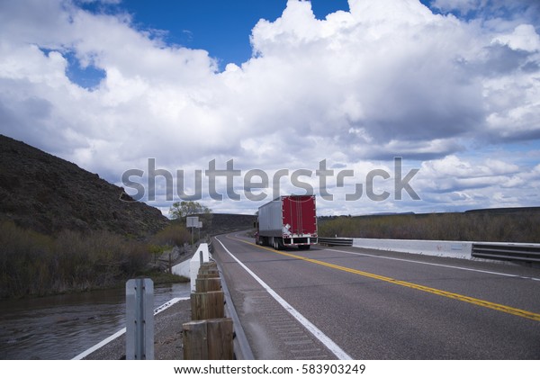 Moving semi truck with the load in\
a dry van trailer with red doors on the divided lanes road pass\
over bridge over a small river that crosses the highway in\
Idaho.