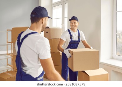 Moving service. Cheerful employees of transportation and delivery company carry cardboard boxes together. Male loaders in overalls are talking cheerfully while packing cardboard boxes in empty office. - Powered by Shutterstock