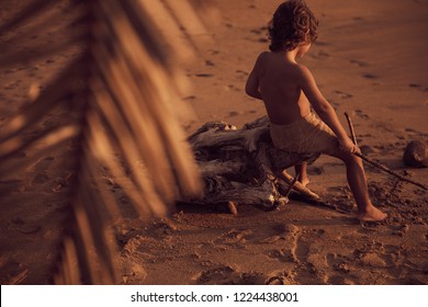 Mowgli boy with curly hair on the beach, sitting on old tree and drawing on sand with a stick. Shot from behind