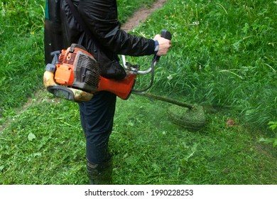 Mows The Grass With A Hand-held Lawn Mower. The Gardener Is Cutting The Lawn. The Gardener's Tool Cuts Tall Grass. The Lawn Mower In Action.