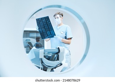 MRI Bore View Of Radiologist In Safety Face Shield Viewing X-ray Image While Analyzing Results Of Computer Tomography