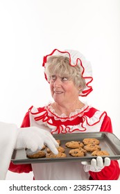 Mrs Clause Looks Up At Santa Who Is Out Side The Frame As Santa's Gloved Hand Is Seen Reaching In To Get A Cookie From Her Tray.