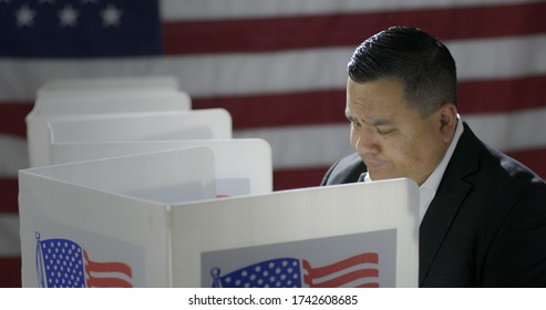 MS Hispanic Man In Polling Station, Voting In A Booth With US Flag In Background. Serious Expression From High Viewpoint