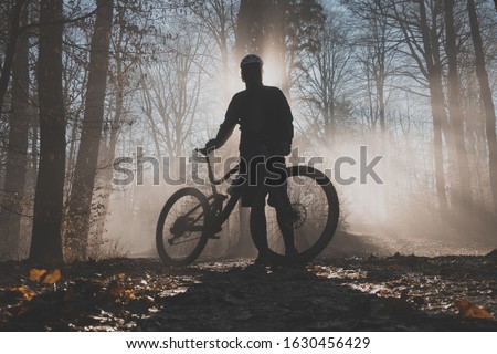 MTB or mountain biker posing in misty forest with sun behind him. Silhouette of a mountain biker in the foggy woods, beautiful sport silhouette