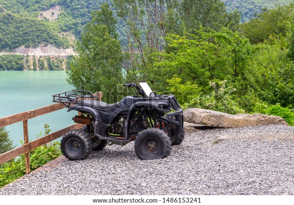 Mtskheta,\
Georgia, 07.20.2019. Black quad bike at the lake in the Parking\
lot, against the green foliage. The Parking lot is covered with\
river pebbles and fenced with a wooden\
fence