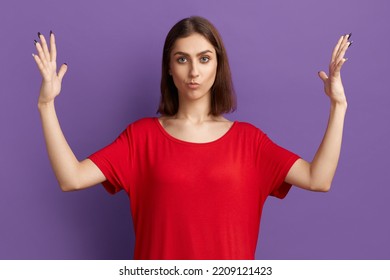 It is too much. Doubting young pretty brunette girl in red t-shirt shows width of something. Woman thinks it is a big price for item. Ask for extra discount. Posing over purple background
