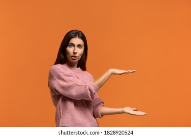 Too Much. Photo Of Sbeautiful Woman Raises Both Palms, Shapes Something Very Big And Wide, Excited With Huge Size, Measures Huge Item, Wears In Pink Knitted Sweater Isolated Over Orange Background