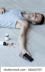 Too Much Pills. Top View Of Young Man Lying On The Floor And Holding A Mobile Phone In His Hand While Bottles With Pills Laying Near Him