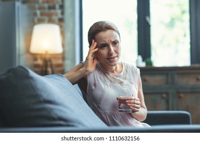 Too Much Stress. Restless Mature Woman Carrying Glass Of Water And Resting On Couch