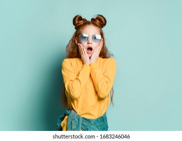 Much Surprised Shocked Red-haired Teenager Girl In Stylish Smoked Blue Glasses And A Yellow Sweatshirt Gasps Covering Her Cheeks With Her Hands