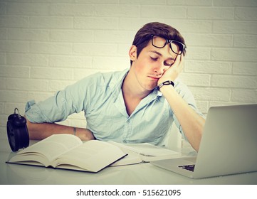 Too much work tired sleepy young man sitting at desk with books in front of laptop computer isolated grey brick wall background. Busy schedule in college, workplace, sleep deprivation concept