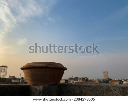 Mud water pot placed on the top of roof wall. Bird water feeder utensil with sunlight and cloudy sky in the background. City: Lahore, Pakistan.