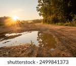 A muddy narrow road with a puddle of water reflecting the trees. Country side forest area. Nature background at sunrise. Nobody. Calm and peaceful mood. Sun flare.