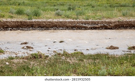 Muddy stream flows through grassy field. Water carries floating debris and sediment. Bank erosion visible along water's edge. Green grass grows on both sides of stream