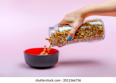 Muesli On A Pink Background. Pour Dry Muesli Flakes By Hand Into A Bowl. Pouring Muesli From A Glass Jar Into A Black Bowl When Making Cereals With Muesli.