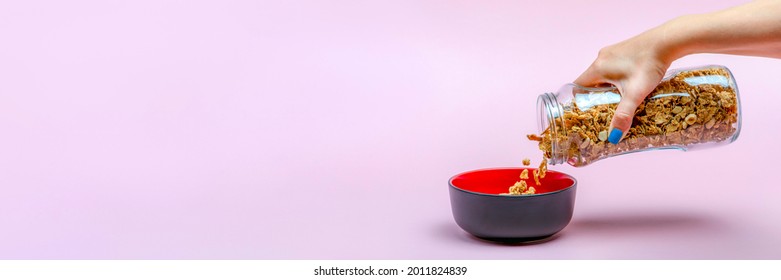 Muesli On A Pink Background. Pour Dry Muesli Flakes By Hand Into A Bowl. Pouring Muesli From A Glass Jar Into A Black Bowl When Making Cereals With Muesli.