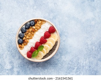 Muesli and yogurt bowl decorated with fresh blueberry, raspberry, banana slices, chia seeds, coconut shred and mint leaf. Healthy vegetarian breakfast or snack. Top view. Copy space. Blue background. - Shutterstock ID 2161541361
