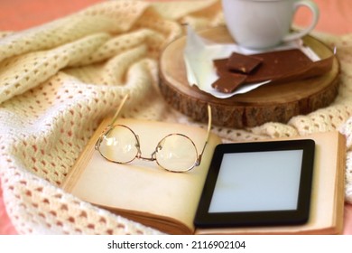 Mug of chocolate chip cookies, bar of chocolate, open book, reading glasses and e-reader on a bed. Selective focus.
