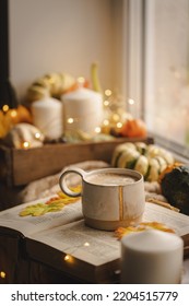 Mug Of Coffee Standing Next To The Window With Candles, Pumpkins, Book And Warm Blanket. Cozy Home Atmosphere In Sunny Autumn Day. Cozy Fall Composition.