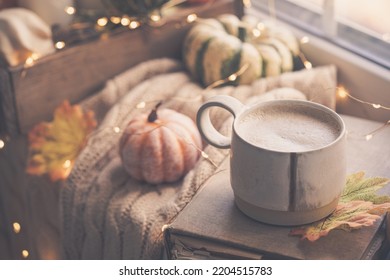 Mug Of Coffee Standing Next To The Window With Candles, Pumpkins, Book And Warm Blanket. Cozy Home Atmosphere In Sunny Autumn Day. Cozy Fall Composition.