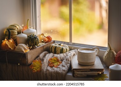 Mug Of Coffee Standing Next To The Window With Candles, Pumpkins, Book And Warm Blanket. Cozy Home Atmosphere In Sunny Autumn Day. Cozy Fall Composition.