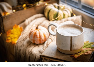 Mug of coffee standing next to the window with candles, pumpkins, book and warm blanket. Cozy home atmosphere in sunny autumn day. Cozy fall composition. - Powered by Shutterstock