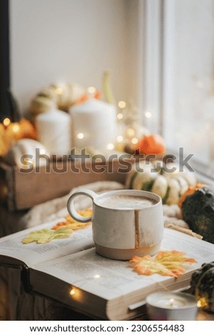 Mug of coffee standing next to the window with candles, pumpkins, book and warm blanket. Cozy home atmosphere in sunny autumn day. Cozy fall composition.