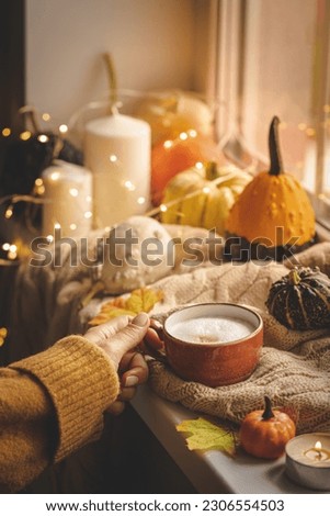 Mug of coffee standing next to the window with candles, pumpkins, book and warm blanket. Cozy home atmosphere in sunny autumn day. Cozy fall composition.