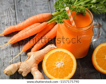 A mug of fresh carrot smoothie with cocktail straw, parsley, ginger root and oranges on a dark wooden table.