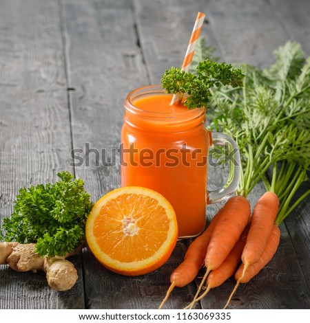 A mug of fresh carrot smoothie with cocktail straw, parsley, carrots, ginger root and oranges on a rustic table.