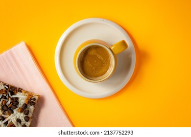 A Mug Of Freshly Brewed Double Espresso Placed On A Saucer In The Middle Of Flat Bright Yellow Background. Layered Cake Decorated With Coconut Pills And Chocolate Chip Placed On Baking Paper.