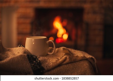 Mug with hot tea standing on a chair with woolen blanket in a cozy living room with fireplace. Cozy winter day. - Shutterstock ID 1838852857