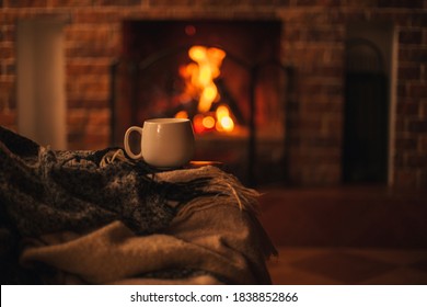 Mug with hot tea standing on a chair with woolen blanket in a cozy living room with fireplace. Cozy winter day. - Shutterstock ID 1838852866
