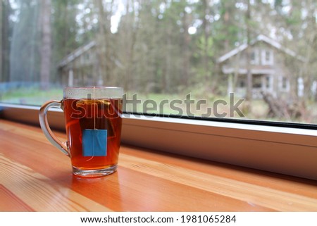 A mug with a tea bag on the wooden windowsill, against the background of pillows with a floral print and a large window with a view of the forest. Tea drinking as a good way to relax or socialize