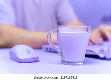 Mug With Unusual Lilac Beverage And A Computer Mouse Are On Table, And Person Who Works Using A Laptop On Blurred Background. Everything Is Painted Purple