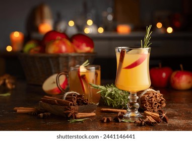 Mulled cider with slice apples, cinnamon, rosemary, and anise stars in glass cups on a brown rustic table. Holiday traditional hot drink. On a dark background kitchen utensils and burning candles. - Powered by Shutterstock