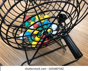 Multi Colored Bingo Ball Numbers In The Selection Cage Sitting On A Wooden Desk.