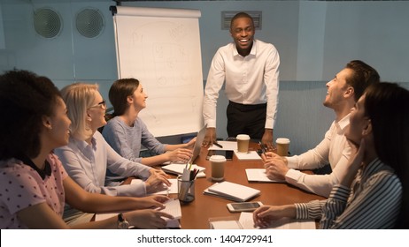 Multi Ethnic Aged And Young Millennial Corporate Team Sitting At Boardroom Listens Looking At Dark Skinned African Team Leader Company Boss Or Business Coach During Evening Meeting At Conference Room