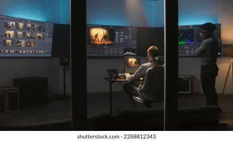 Multi ethnic colorist and film maker edit video and make color grading in program. Process of colour correction for movie post production in modern studio. Multiple monitors with action film footage.