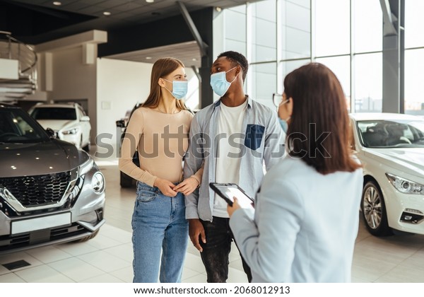 Multi ethnic\
couple in face mask talking with female dealer while choosing new\
auto at showroom. Competent manager with tablet in hands providing\
consultation for clients.