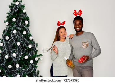 Multi ethnic couple posing next to Christmas tree. White wall in background and champagne, present and wine glasses in hands