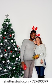 Multi ethnic couple posing next to Christmas tree. White wall in background and champagne, present and wine glasses in hands