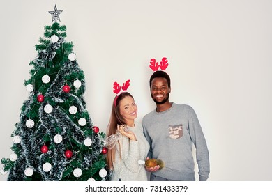     Multi ethnic couple posing next to Christmas tree. White wall in background and champagne, present and wine glasses in hands 